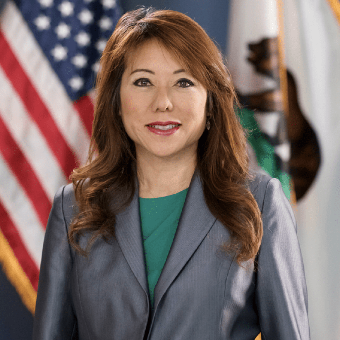 A woman in a gray blazer and green top stands in front of American and California flags.